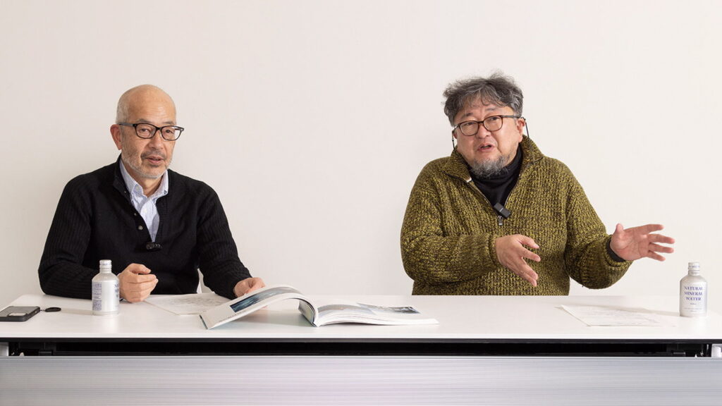 眼鏡をかけた二人の男性が白いテーブルを挟んで座り、向かって右側の男性が身振り手振りを交えて話し、左側の男性が手元の開かれた冊子を見ながら話を聞いている様子を捉えた屋内での対談シーンの写真