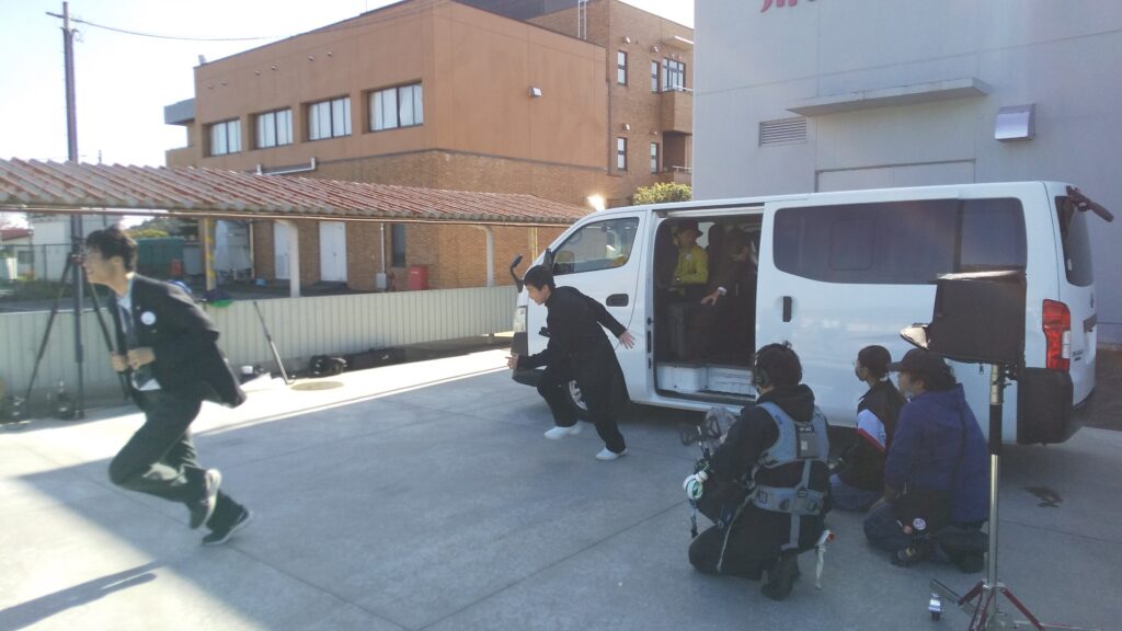 白いワゴン車の横で走り出す人物を周囲のスタッフが機材を構えて撮影している様子を捉えた写真