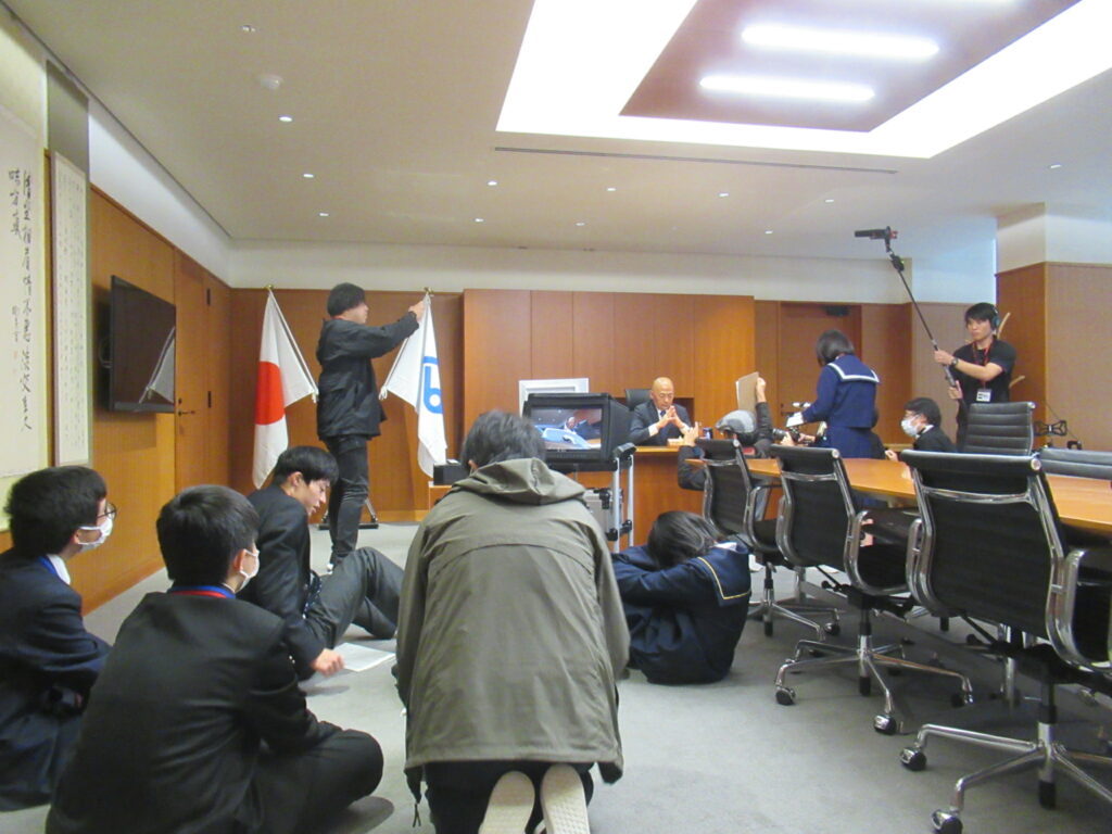 会議室でスタッフや学生たちが旗や機材を用いて撮影の準備と段取り確認を進める様子を捉えた写真