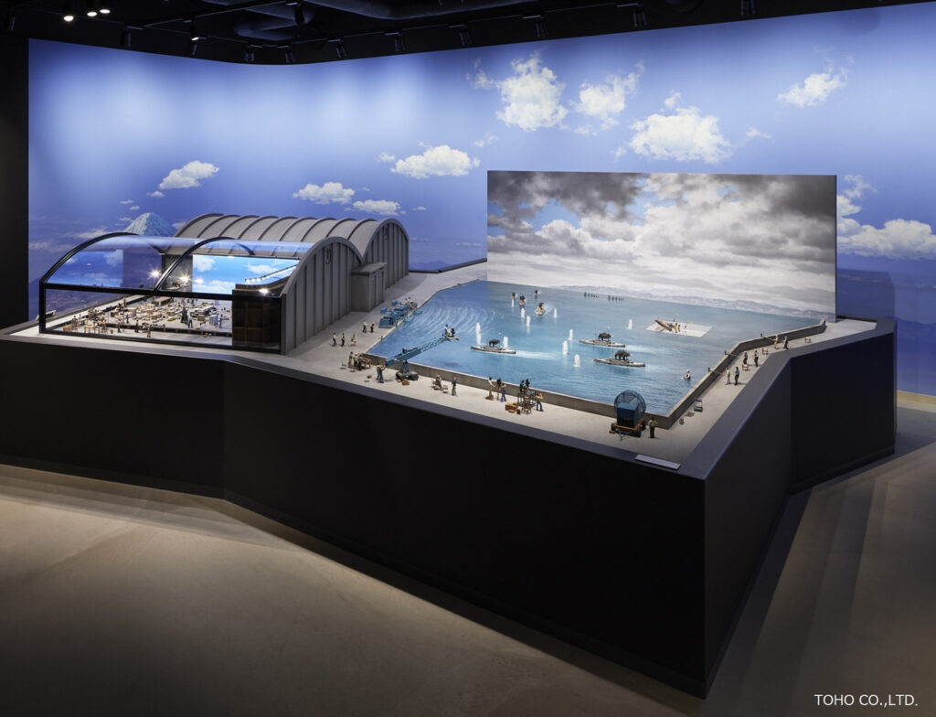 大規模な海のミニチュアセットで、水面に浮かぶ艦船模型や遠景の青空、山々が描かれた背景画などのセットの写真