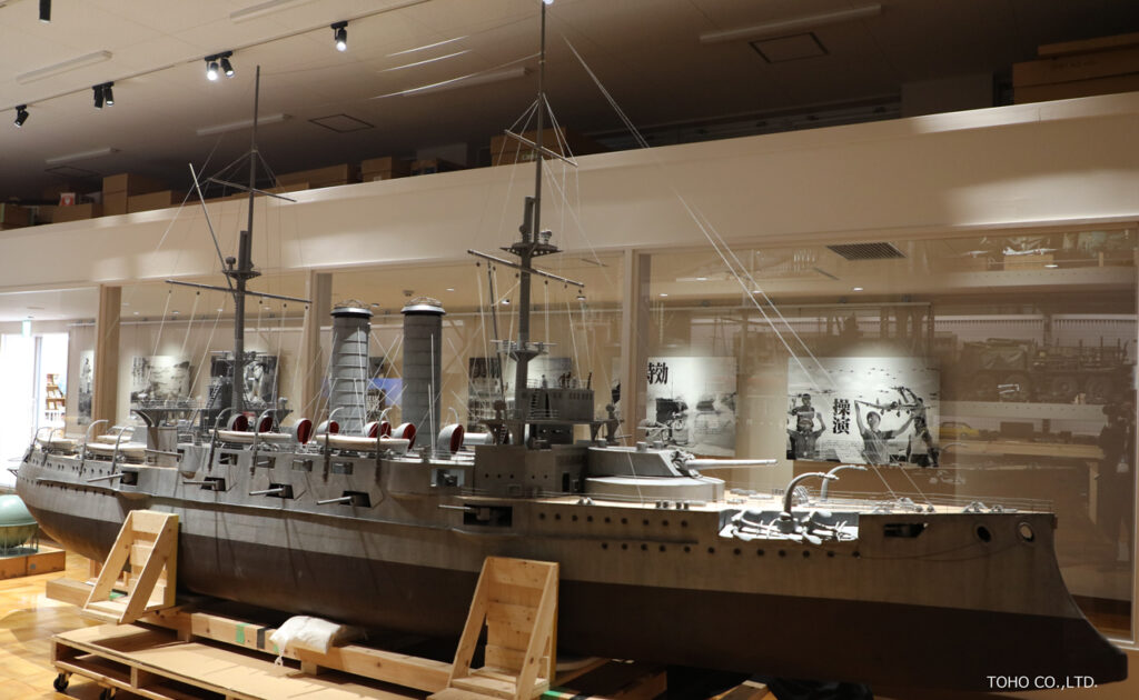 巨大な精密なミニチュア模型「戦艦三笠」が木製の台座に安置され、背景には特撮映画の撮影風景を記録したパネルや機材が並ぶ展示室の写真