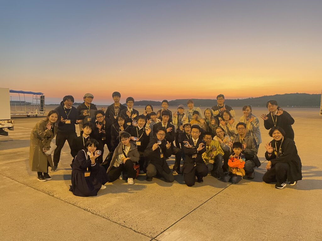 夕暮れの滑走路でたくさんの関係者が寄り添い明るいポーズを取り柔らかな空の色に包まれた雰囲気を伝える写真