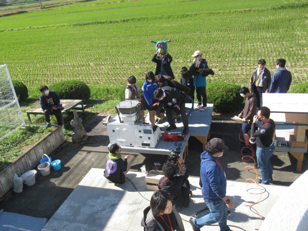 のどかな田園風景を背景にした屋外の撮影現場で、飛行場の管制塔のような白い建物模型の周りに学生たちが集まり、怪獣の頭部プロップを掲げたりカメラを構えたりしながら、特撮シーンのセッティングを協力して進めている風景の写真