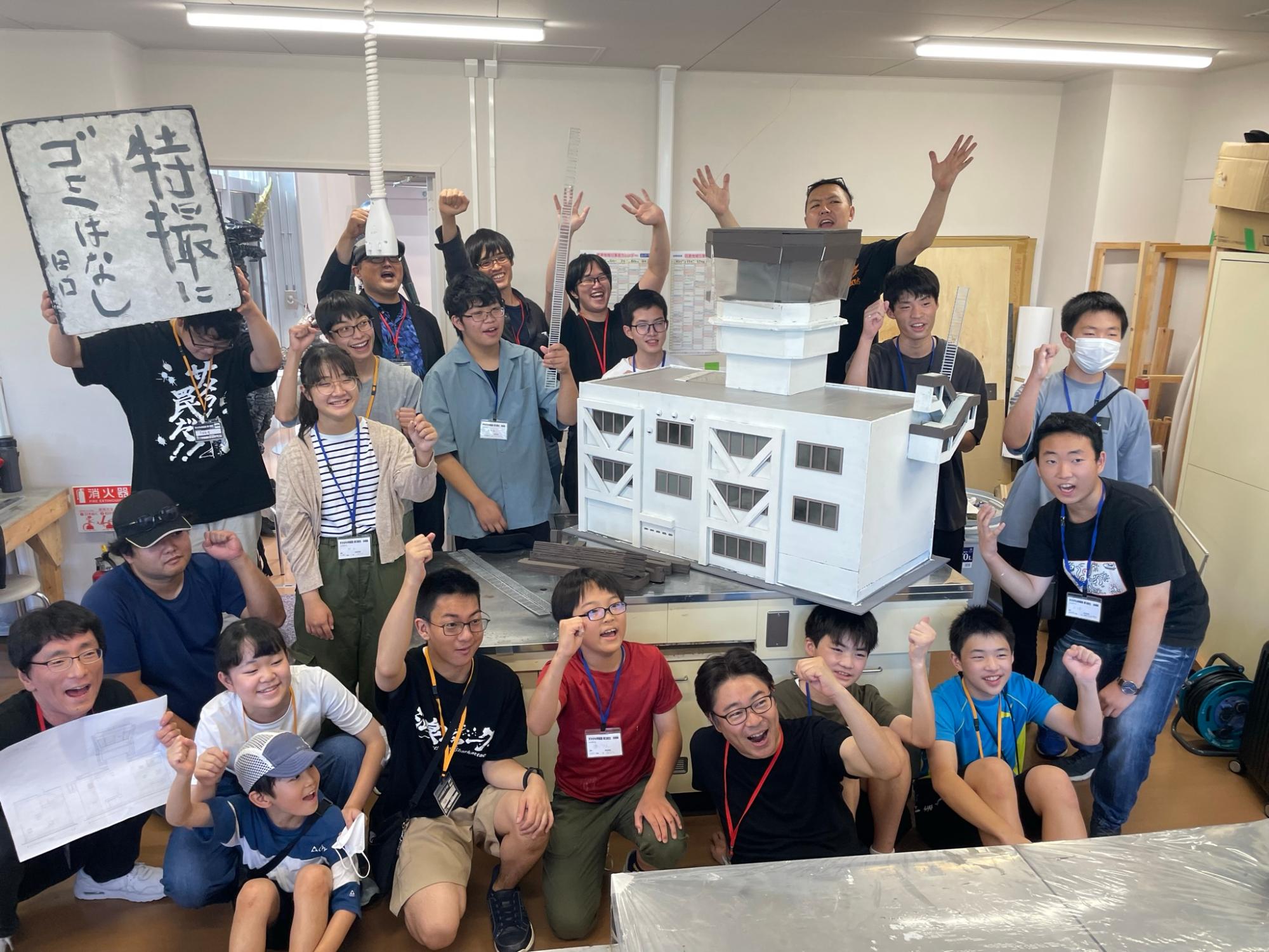 室内で大きな白い建物の模型を囲み、笑顔で拳を突き上げたり手を広げたりして達成感を表現している多世代の男女グループの塾生と、左上で「特撮にゴミはなし」と書かれた看板を掲げる男性を捉えた集合写真