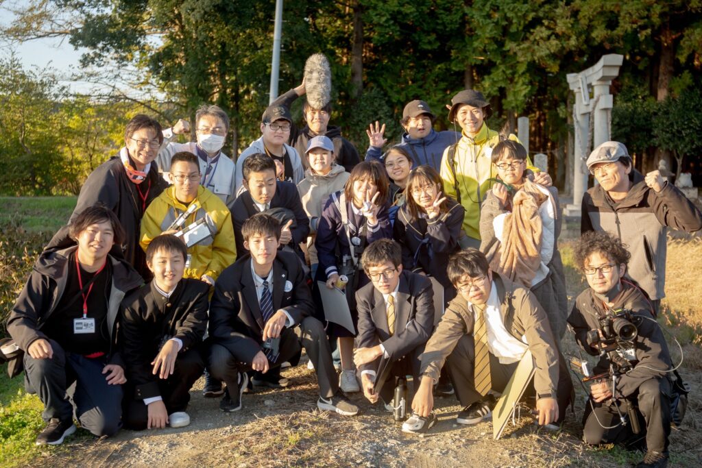 夕暮れ時の柔らかな光が差し込む屋外で、制服姿の学生たちやカメラ・音声機材を手にしたスタッフたちが、充実感あふれる表情で肩を寄せ合い集合写真に収まっている、特撮ワークショップでの写真