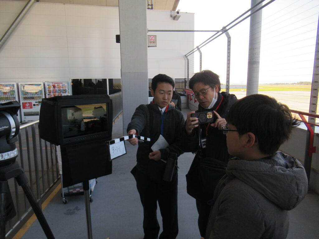 屋外の通路でモニターとカメラの前に集まった人々がクラッパーを構えて映像を確認しつつ撮影準備を進める写真