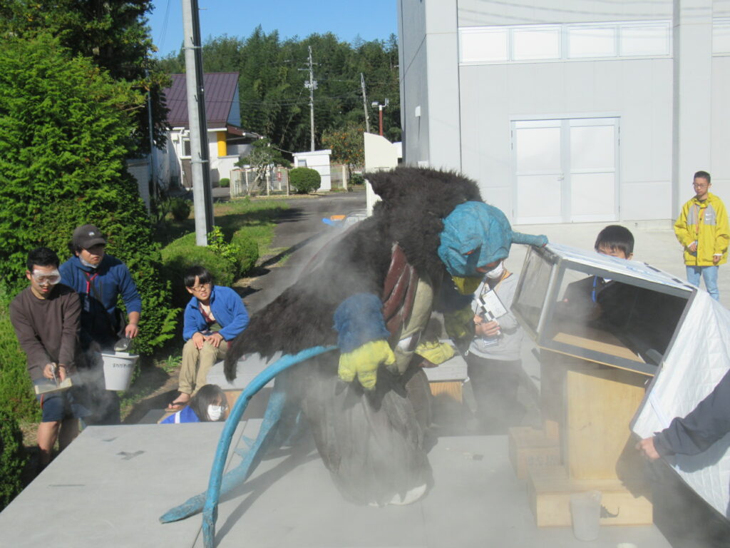 屋外の撮影現場で、大きな一つ目の怪獣スーツが白い煙に包まれながら精巧な建物模型を破壊するシーンを、学生たちがスモークマシンやブロワーを駆使して演出している、特撮映像制作ワークショップの熱気あふれる風景を捉えた写真
