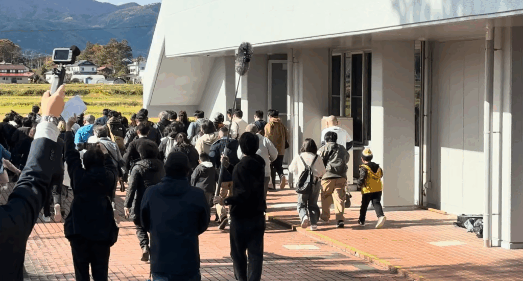 屋外の建物横を大勢の人々が同じ方向へ歩いており、その様子を撮影スタッフがカメラやブームマイクを構えて広報から撮影している場面の写真