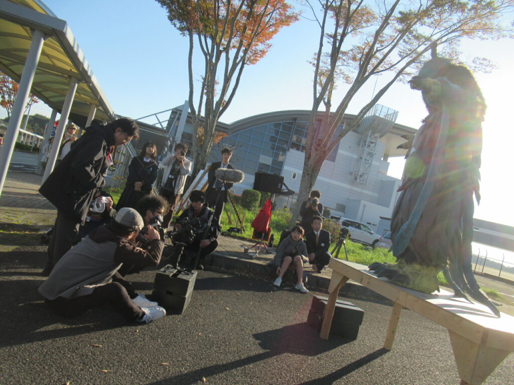 秋の陽光が降り注ぐ屋外の撮影現場で、台座の上に立つ大きな怪獣スーツを正面に据え、カメラマンや録音スタッフ、そしてそれを見守る学生たちが真剣な表情で撮影に臨んでいる、特撮映画の制作風景を捉えた写真