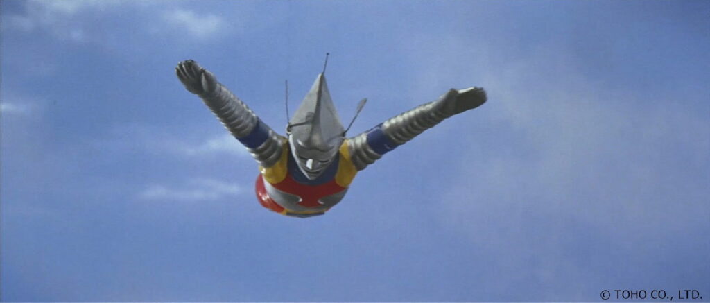 青空をバックに特撮ヒーロー「ジェットジャガー」が両腕を斜め上に大きく広げ、銀色のマスクと蛇腹状の腕を輝かせながら、ダイナミックに飛行している劇中のワンシーンを捉えた特撮写真