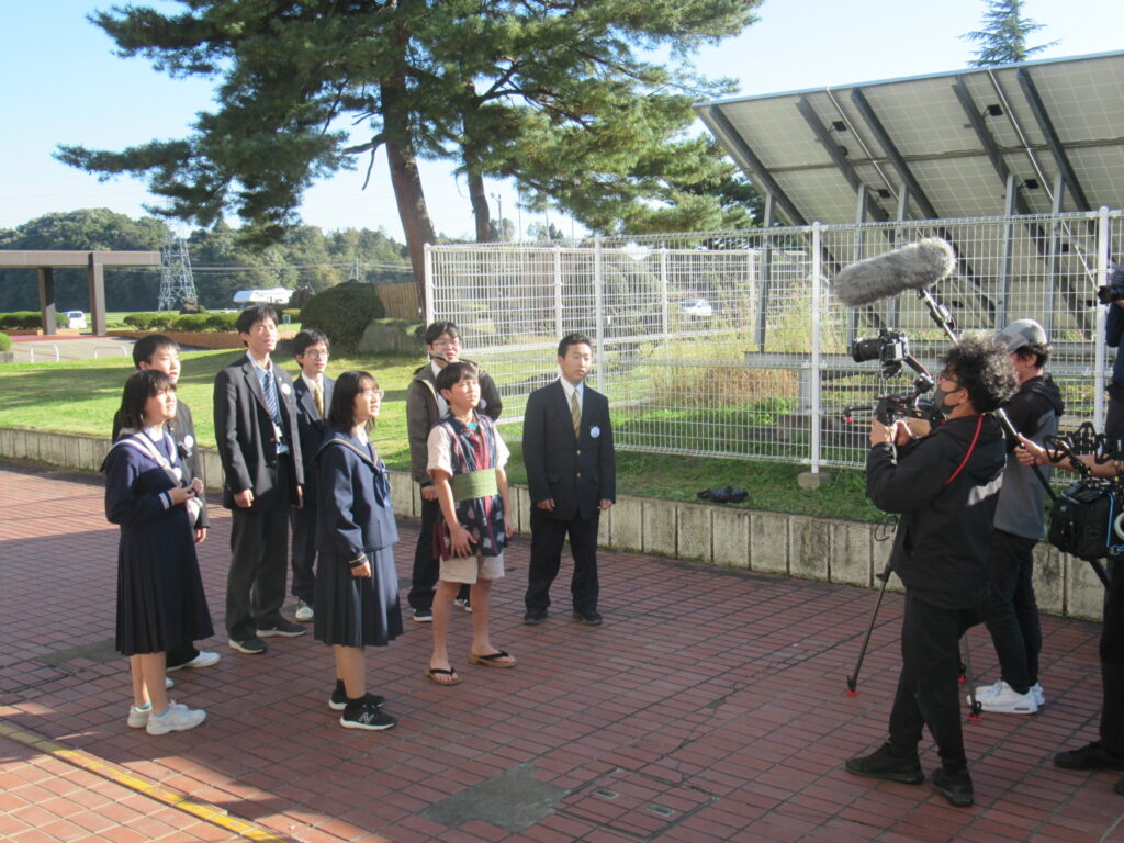屋外の明るい広場で数人の学生が前方を見つめて整列し撮影スタッフが機材を構えて収録する写真