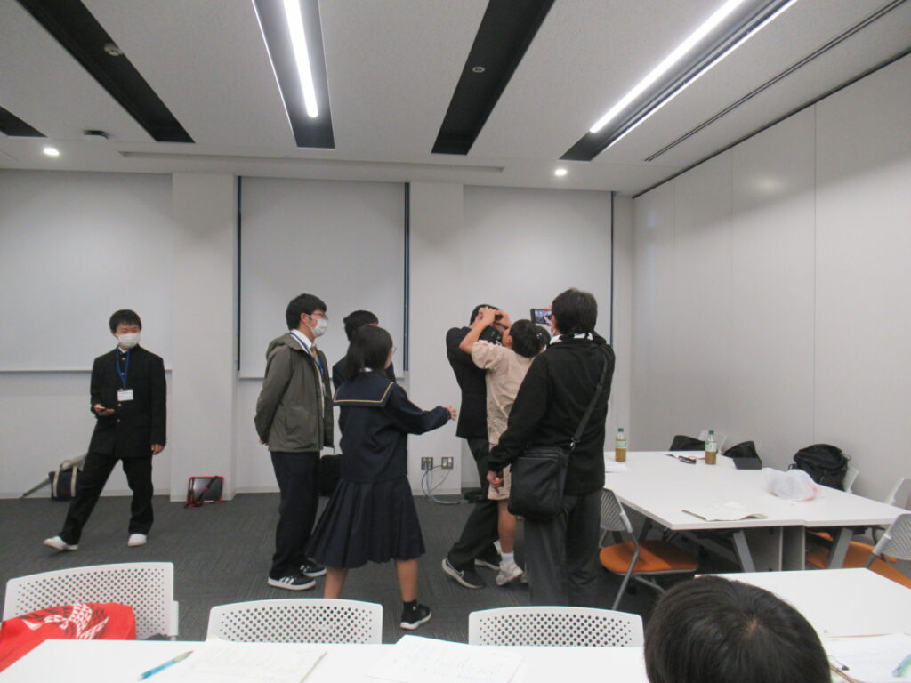 白を基調とした会議室で、制服姿の女子学生や私服の男子学生たちが一箇所に集まり、スマートフォンの画面を覗き込んだりポーズを相談したりしながら、和気あいあいとした雰囲気で撮影の準備や交流を行っている風景の写真