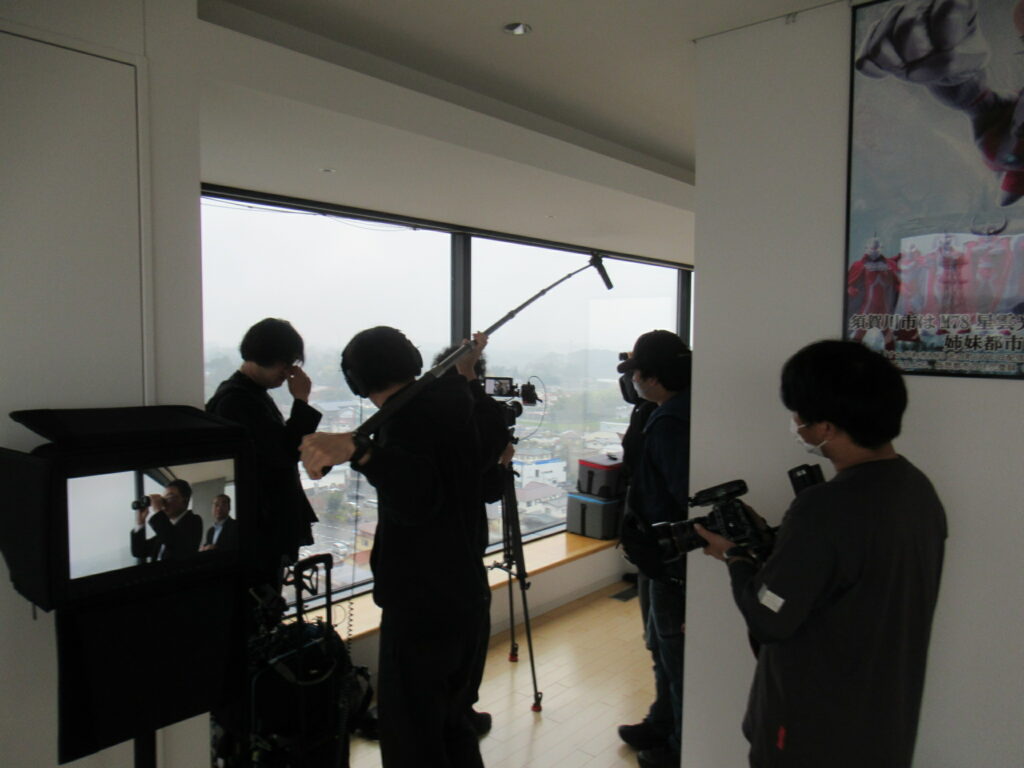 大きな窓から街並みを望む室内で撮影機材を構えたスタッフたちが照明や音声を調整しながら撮影準備を進めている写真