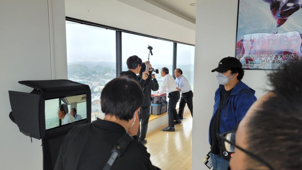 大きな窓のある室内で撮影スタッフがモニターや音声機材を操作しながら人物の演技を撮影している様子を捉えた写真