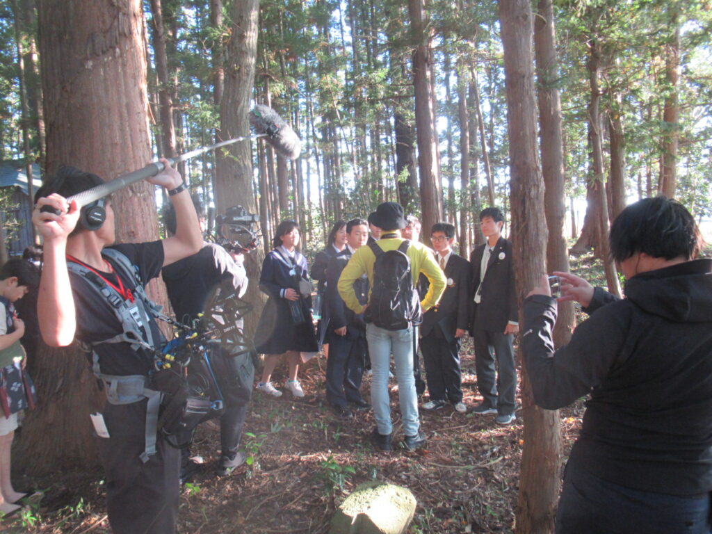 木立から柔らかな光が差し込む森の中で学生らが話し合う様子を撮影スタッフが周囲で機材を構えて丁寧に収録する写真