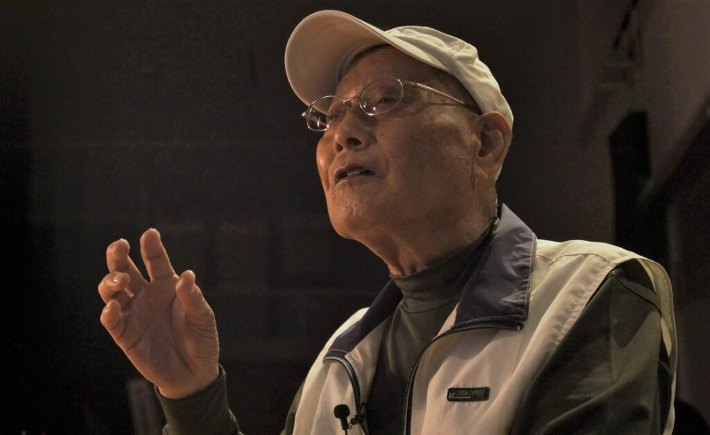 白いキャップを被り眼鏡を掛けた年配の男性が、暗い背景の中で右手を軽く上げながら、穏やかな表情で斜め上を見つめて何かを熱心に語っているような、胸から上を捉えたポートレート写真