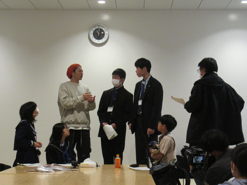 室内で時計の下に集まった人々が資料を手に説明を交わし、周囲のメンバーがその様子を見守る場面を捉えた写真