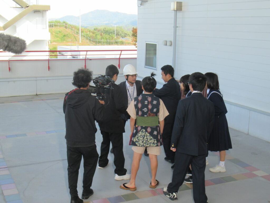屋外のテラスで学生たちが白いヘルメットの人物を囲み撮影スタッフが機材を構えて収録する写真