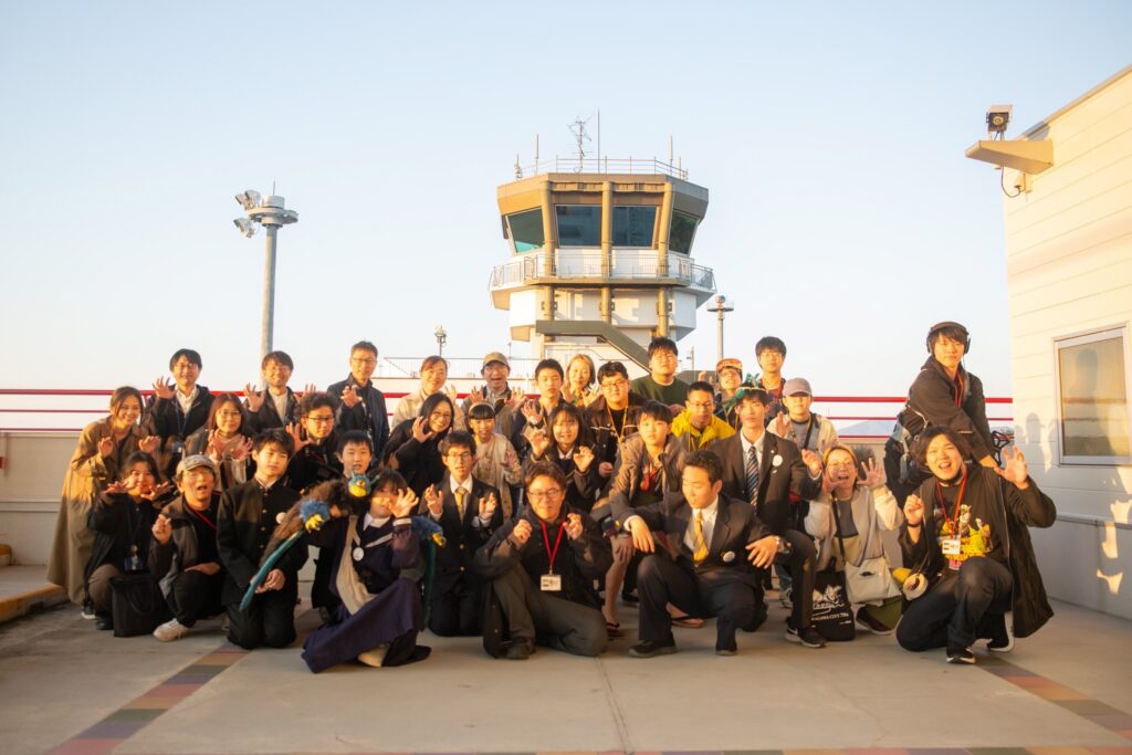 夕陽に照らされた管制塔前でたくさんの関係者が肩を寄せ合い賑やかにポーズをとる様子を収めた写真