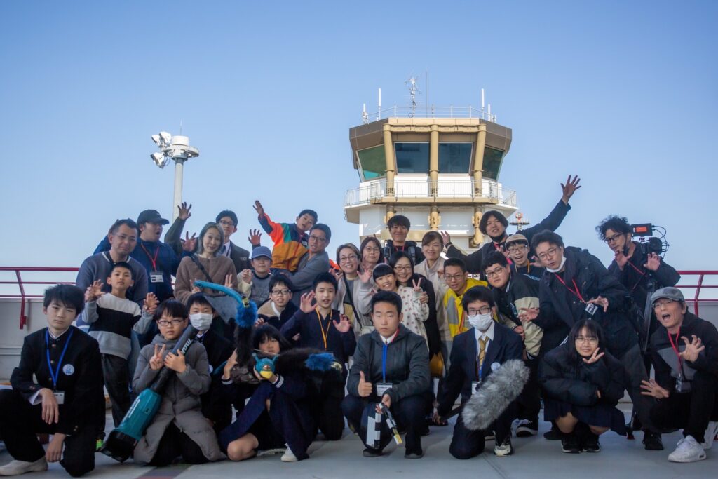 青空の下で管制塔を背景に大人数が手を広げポーズを揃えてにぎやかに集合する様子を捉えた写真