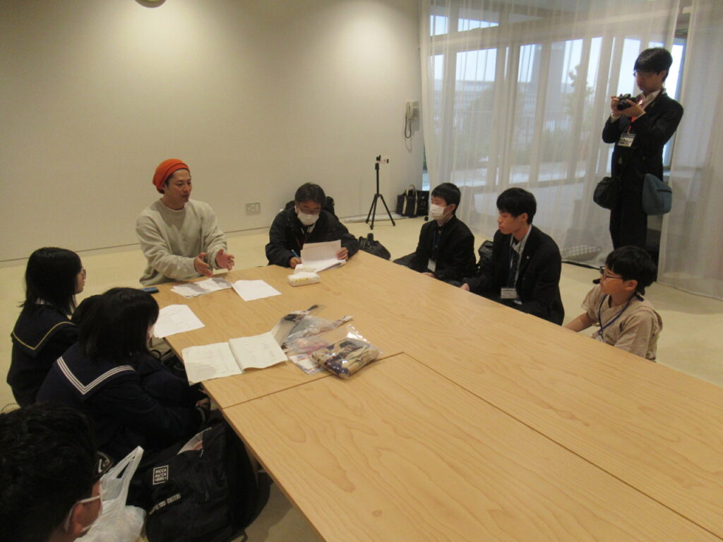 室内の大きな木製テーブルを囲んで人々が資料を手に話し合い、後方では別の人物がその様子を記録している写真