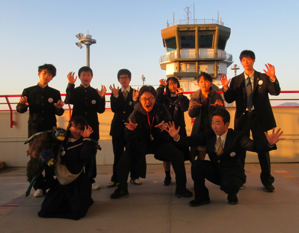 夕陽に照らされた管制塔前で制服姿の若者たちが手を広げてポーズを取り活気ある雰囲気を伝える写真