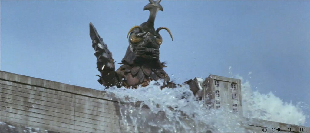 カブトムシのような大きな角とドリル状の腕を持つ怪獣メガロが、激しい水しぶきを上げながらダムを破壊して出現している迫力あるワンシーンを捉えた写真
