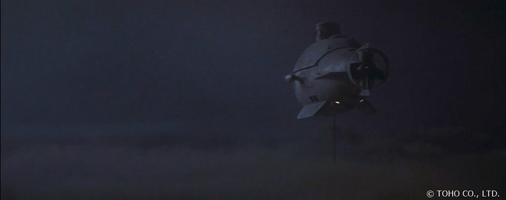 暗い海中を模した空間で、潜水艇「わだつみ」がサーチライトを点灯させながら静かに潜航している様子を後ろから捉えた写真