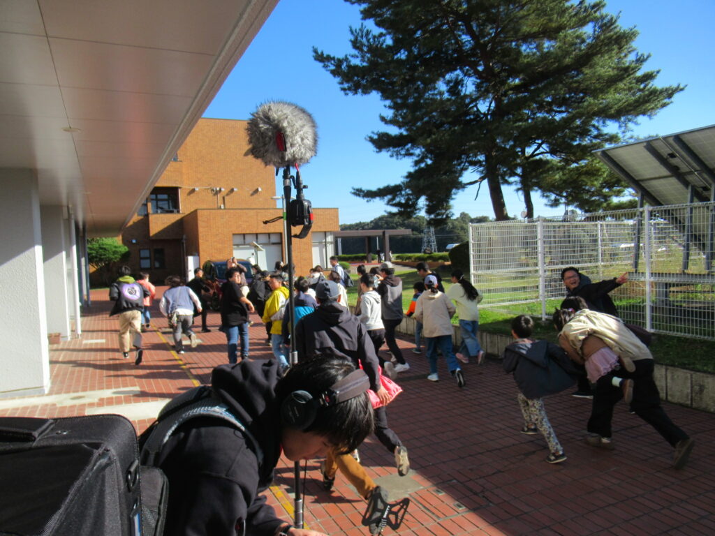 青空の下の建物脇で多くの人々が一斉に駆け出す様子を撮影スタッフが機材越しに追う写真