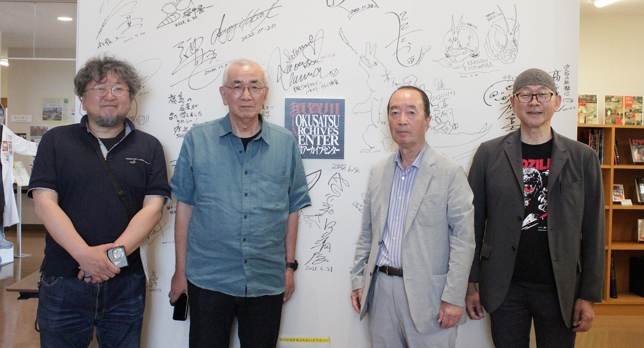 サインが書き込まれた白いバックパネルを背にして、四人の男性が横一列に並び、カメラを見つめて記念撮影を行っている写真