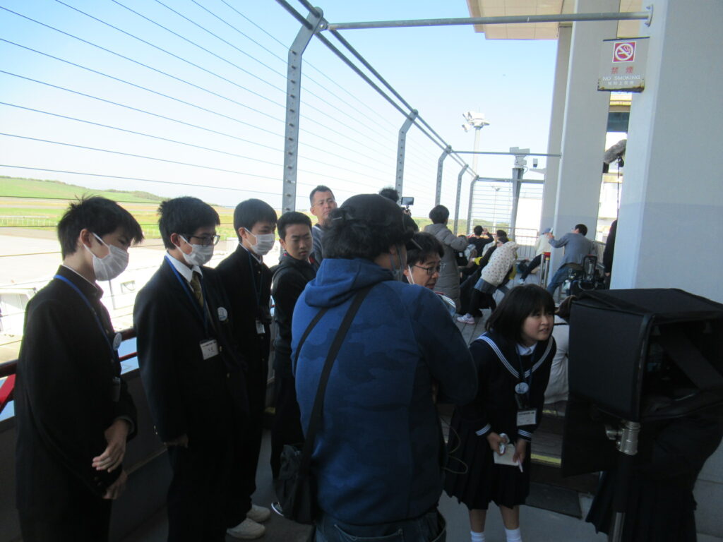 屋外のフェンス沿いで学生たちが撮影スタッフと共に機材を前に並び撮影準備を進めている写真