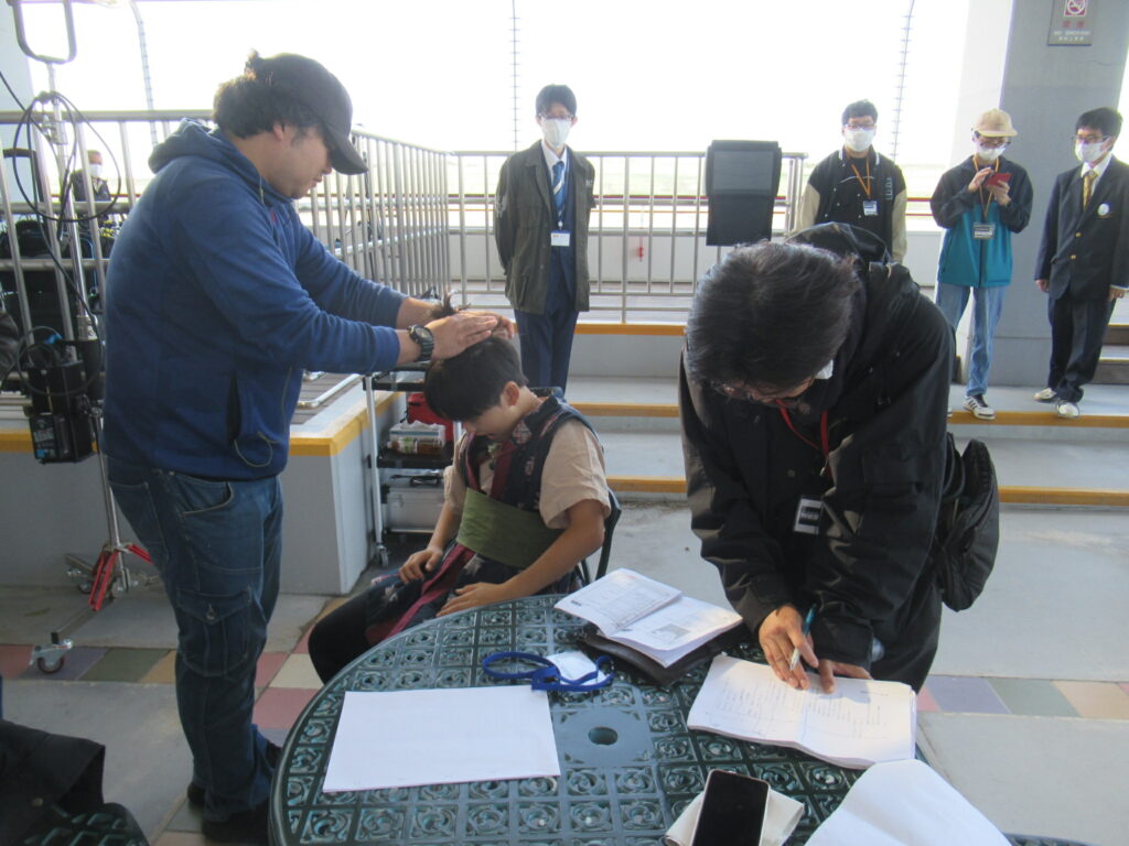 屋外のテラスでスタッフが椅子に座る人物の頭部を整え、別のスタッフが書類を確認しながら準備を進める写真