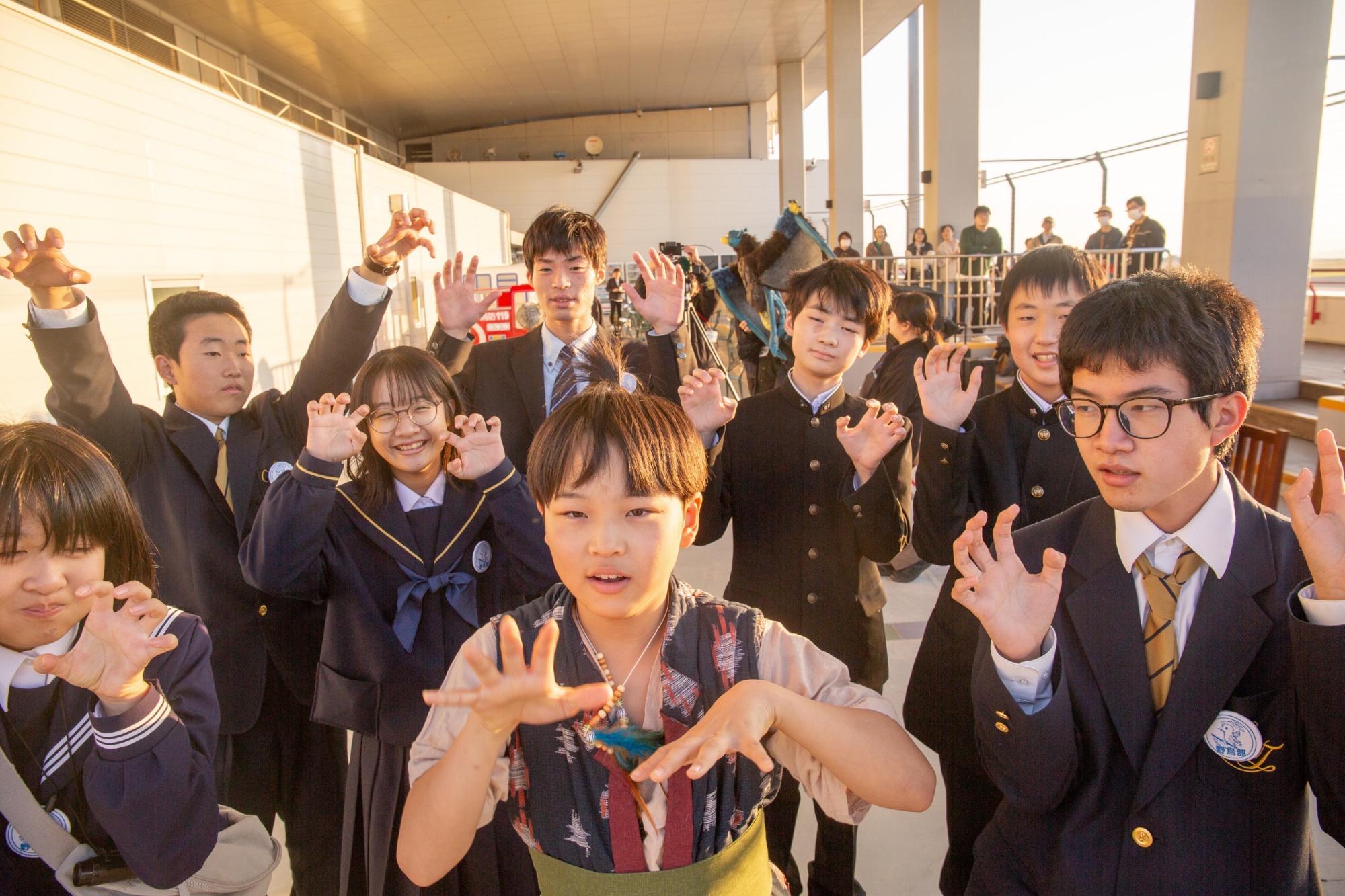 夕日の差し込む屋外通路で制服姿の若者たちが両手を上げ怪獣のようなポーズを揃えて楽しげに構える写真