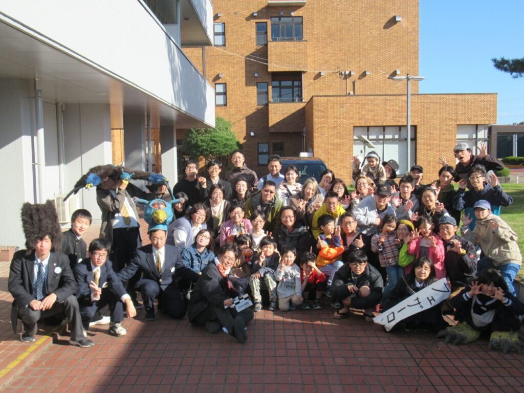 日差しの下、建物前に大勢の人々が怪獣の着ぐるみも交えて笑顔で賑やかに記念撮影する写真