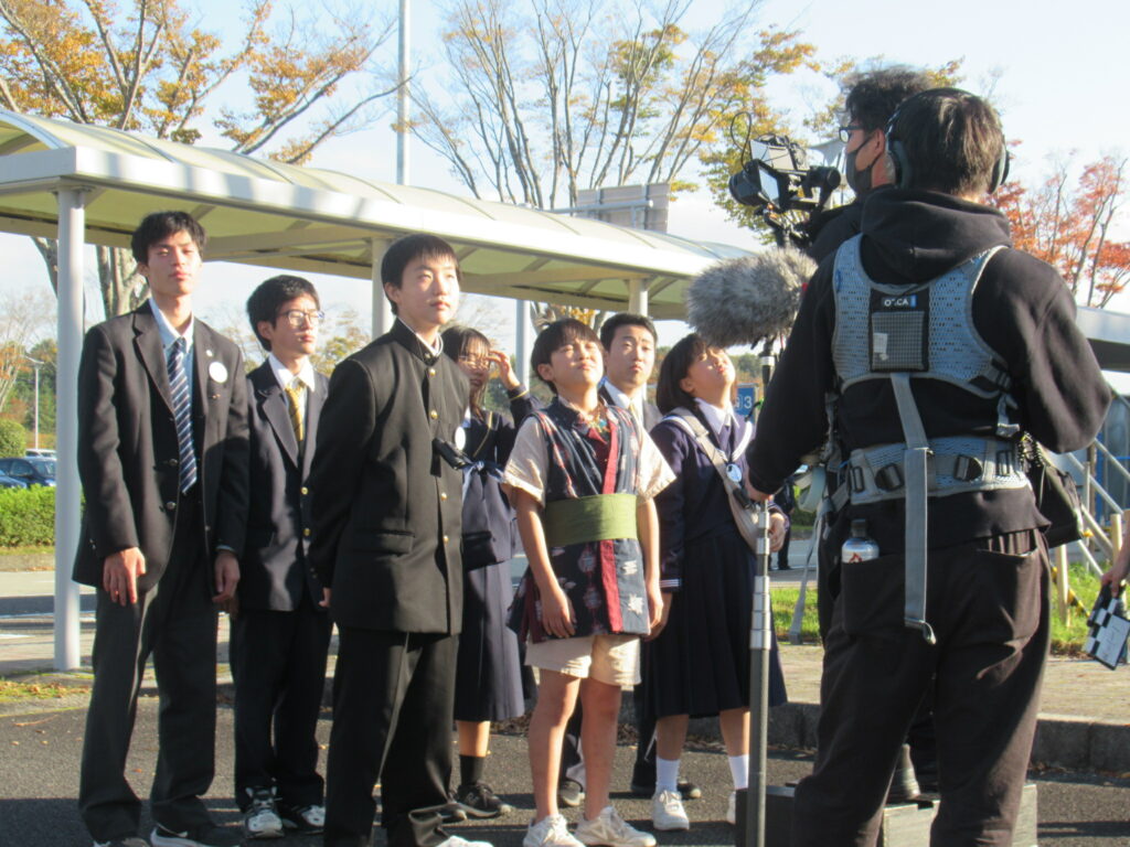 屋外のバス停付近で制服姿の学生たちが並び、撮影機材を構えたスタッフが前方から取材する様子の写真