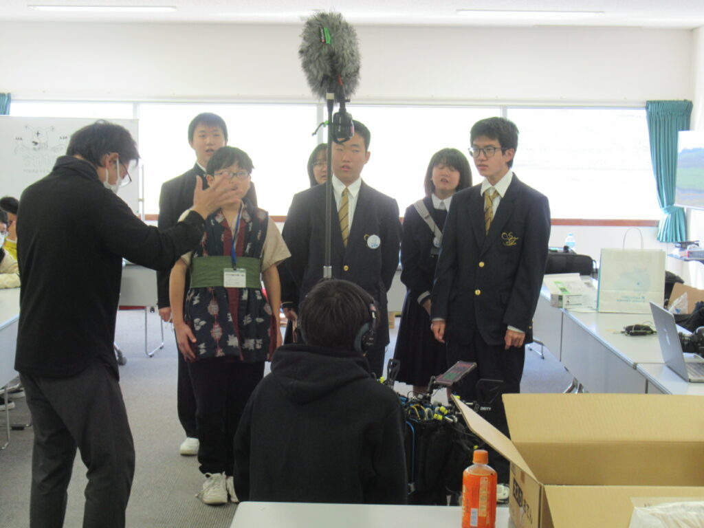 室内で学生たちが並んで撮影に臨みスタッフが指示を出し周囲に録音機材が配置された様子を捉えた写真