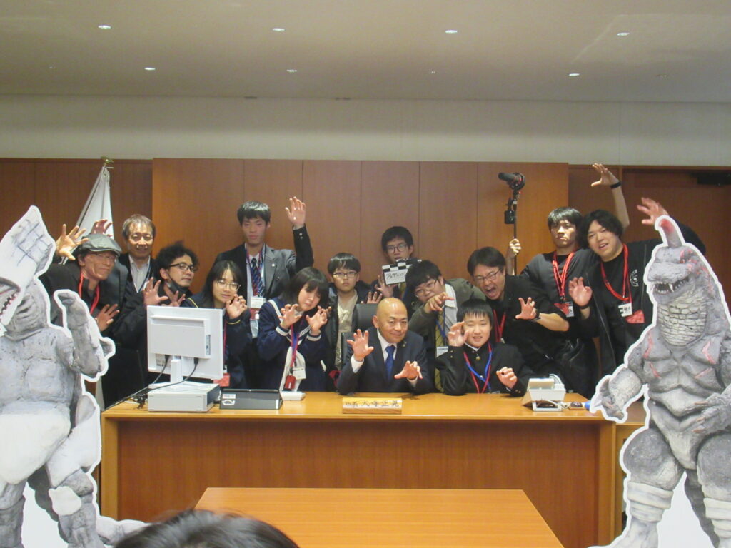 木製机の前に集まった大勢が怪獣パネルに囲まれながら手を上げて賑やかにポーズを揃え記念撮影する写真