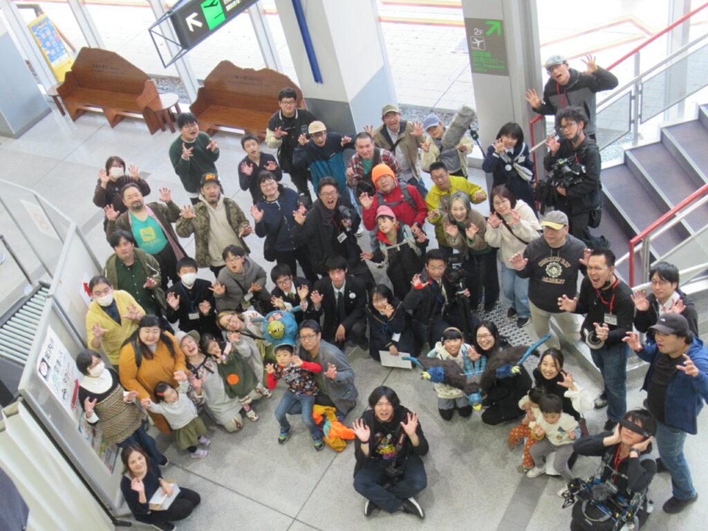 駅構内の広いホールで大勢の人々が階段付近に集まり手を振って和やかに記念撮影に臨む様子を上方から収めた写真