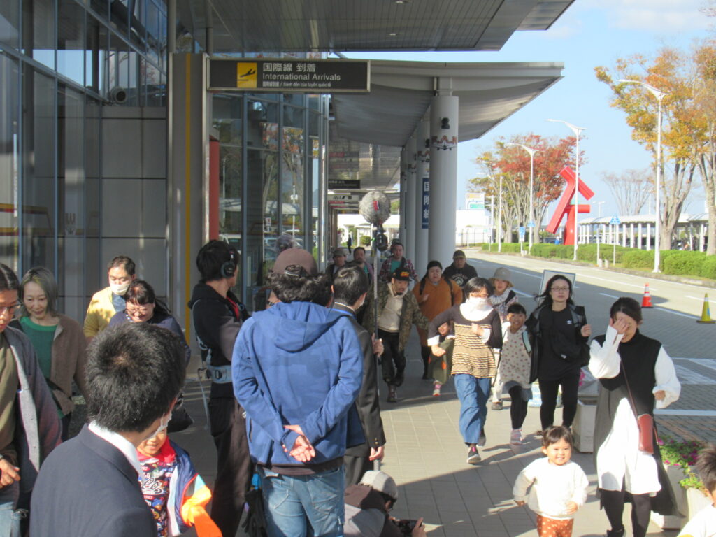空港の国際線到着口前で多くのエキストラが歩道を行き交う中、秋色の木々と近代的な建物が並ぶ風景を撮った写真