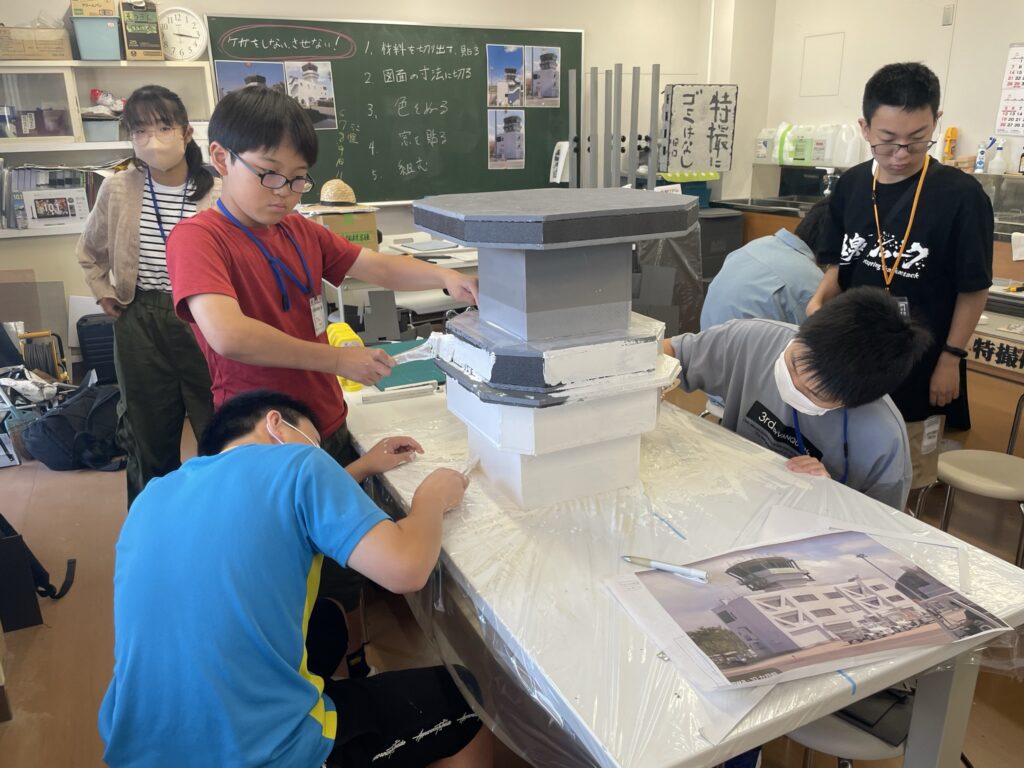 作業室内で塾生が、白いビニールで保護された机の上に置かれた複雑な多角形の塔のような模型に、刷毛を使って白い塗料を丁寧に塗り重ねている作業風景の写真