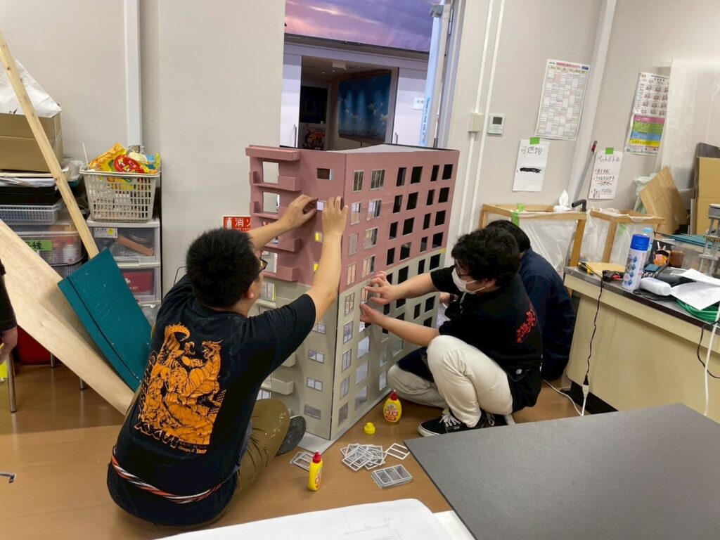室内で二人が床上に据えた大型の建物模型に窓パーツを接着しながら細部を調整して組み立て作業を進める写真