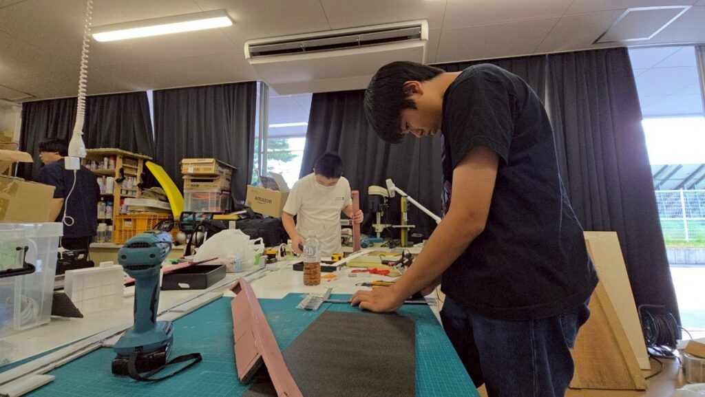 明るい実習室で、手前にいる黒いTシャツ姿の男子学生がカッターと定規を使って黒いボードを丁寧に切り出しており、奥でも白いTシャツの学生が別の部材を加工するなど、各自が集中して模型制作に取り組んでいる風景を捉えた写真