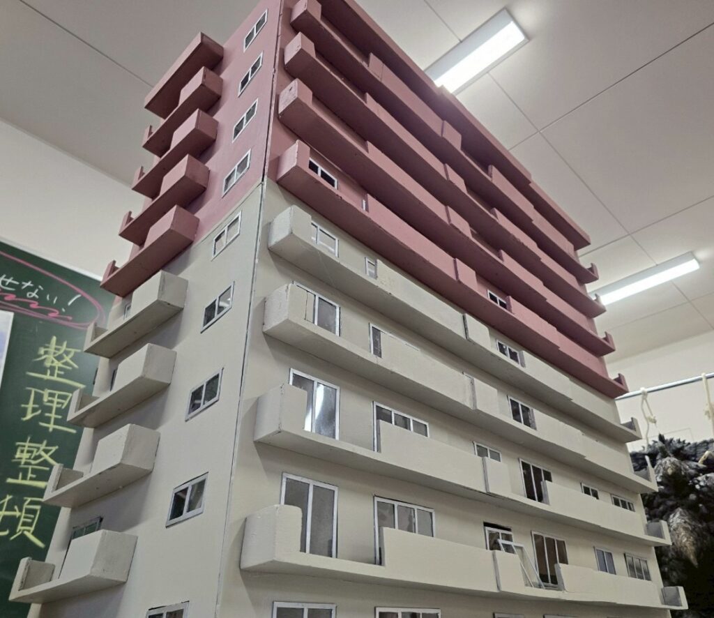 室内で撮影されたピンクと白の外壁と多数のバルコニーを備えた高層マンションの精巧な建築模型を下から見上げる写真