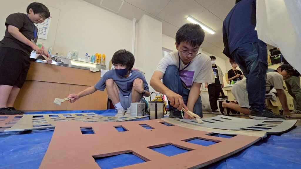 青いシートが敷かれた実習室の床にしゃがみ込み、窓の形に切り抜かれた大きな建物の模型パーツに対して、刷毛を使いながら塗料を丁寧に塗り進めている学生たちの真剣な制作風景を捉えた写真