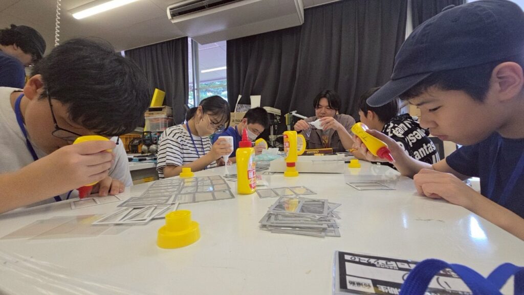 明るい実習室の中で、学生たちが白い作業テーブルを囲み、木工用接着剤を使って建物の窓枠のような小さなシルバーの部材を一つずつ丁寧に貼り合わせている、集中した空気感が伝わる模型制作風景の写真