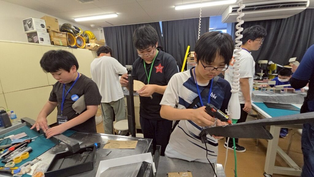 作業室で塾生たちが道具を使い黒い部材の切断や接着作業を行いながら模型制作を進めている写真
