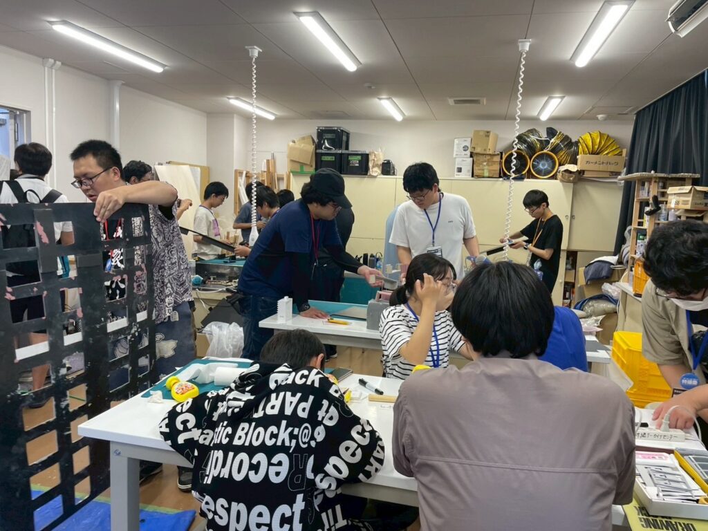 明るい実習室の中で、学生たちが複数のテーブルに分かれて模型制作に励んでおり、手前では建物の窓枠のような黒い部材を調整したり、白やボーダーの服を着た学生たちが真剣な表情で作業を分担したりしている制作風景の写真