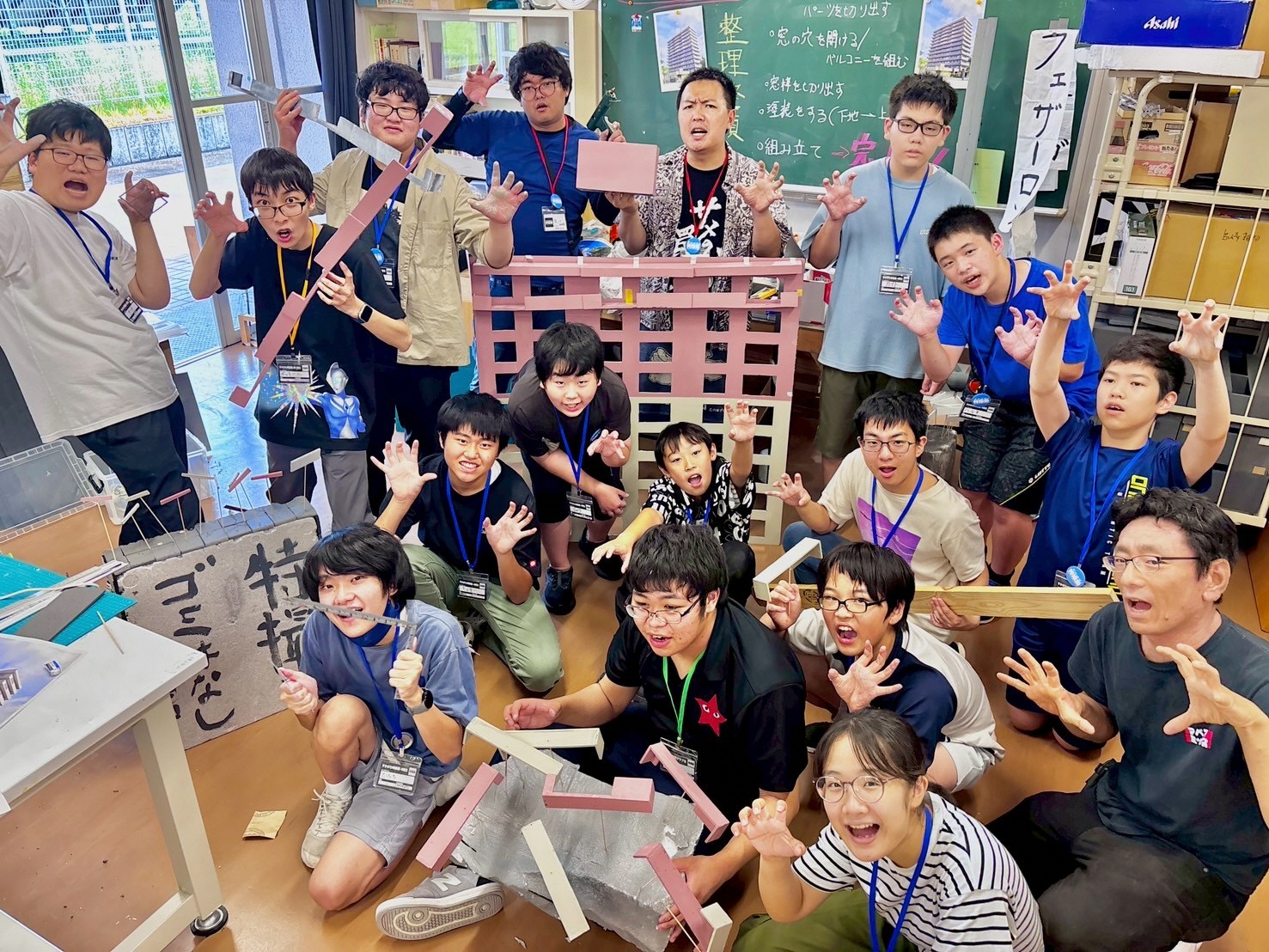 教室のような場所で、制作途中の大きなピンク色の建物の模型や工作部材を囲み、多くの学生たちが怪獣のように両手を広げるポーズをとりながら、賑やかで楽しそうな表情で集合写真に収まっている様子を捉えた写真