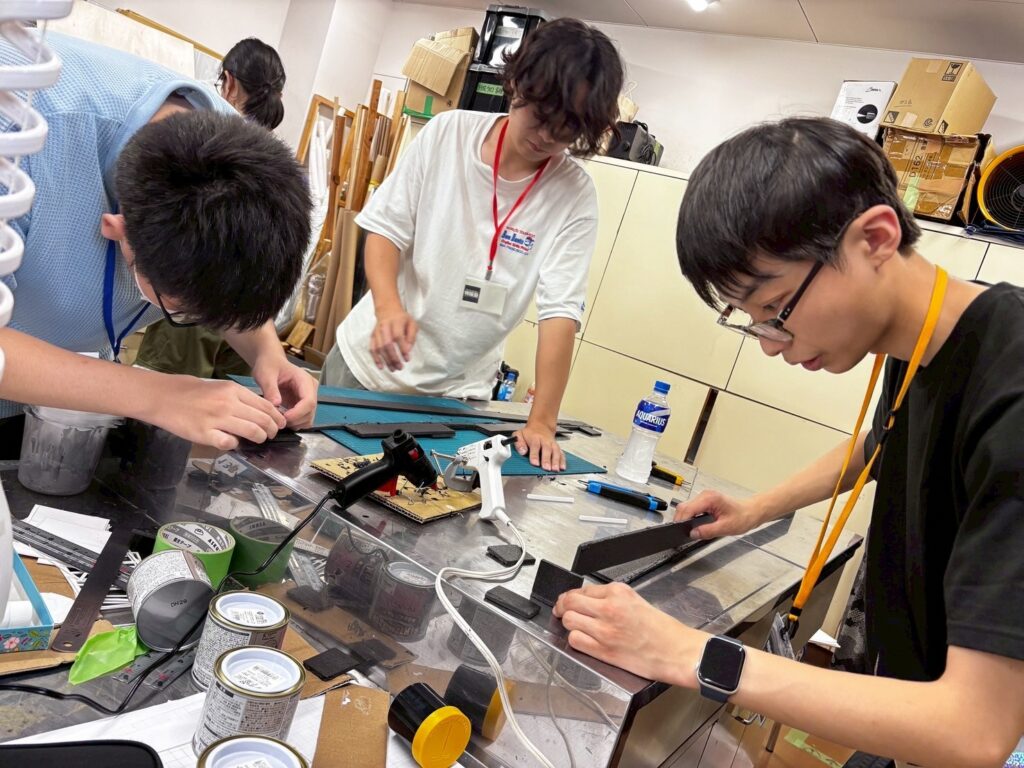 作業室で複数の塾生がカッターマット上で部材を切り出したり接着作業を進める様子を捉えた写真