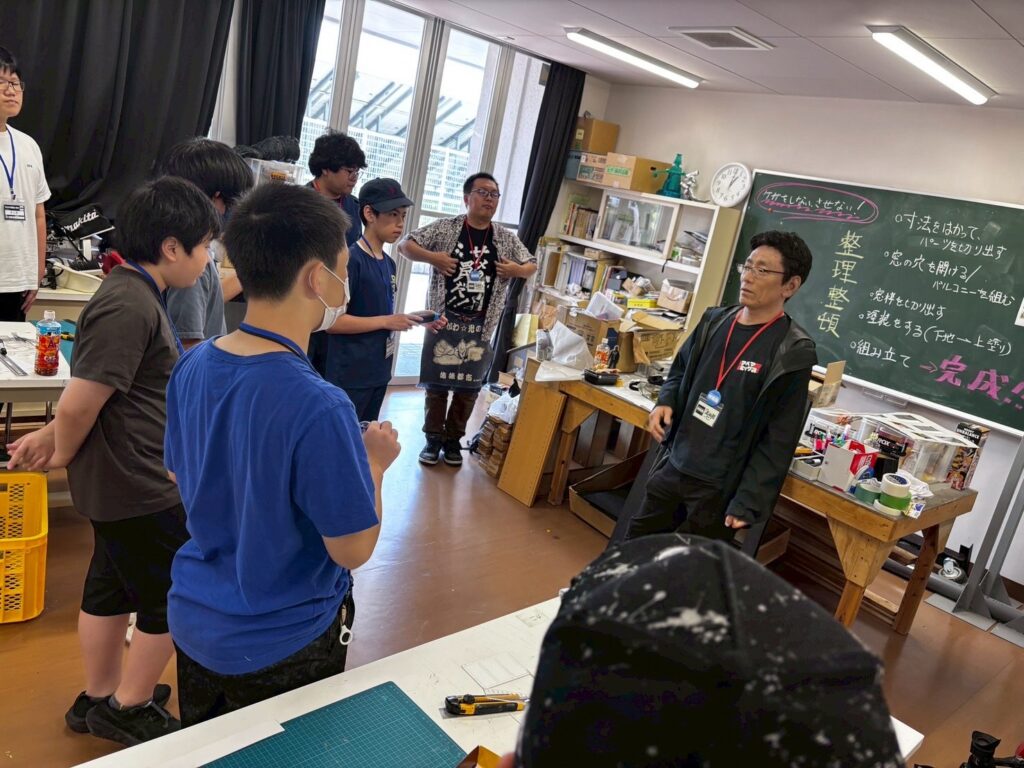 制作工程が書かれた黒板のある実習室で、黒い上着を着た講師が学生たちに向かって説明を行い、周囲には工作道具や資材が並ぶ中で学生たちが真剣な表情で話を聞いている写真